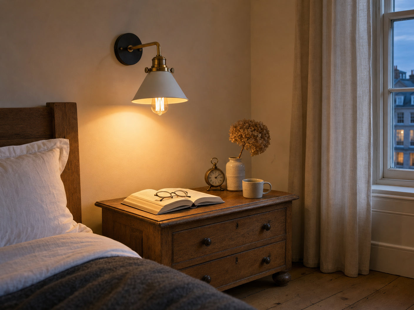 Bayswater white cone wall sconce with brass collar mounted in a calm British townhouse bedroom above a low oak nightstand at headboard height, pooling warm Edison glow onto an open hardback book, reading glasses, brass alarm clock, and a small ceramic vase of dried hydrangea, sash window onto blue-hour twilight