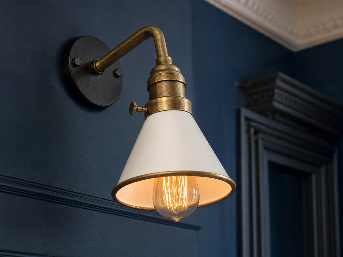 Close-up product detail of Bayswater wall sconce showing the antique-brass collar with horizontal knurled thumbscrew, brass swing-arm, white-painted metal cone shade with brass rim, glowing Edison filament bulb, mounted on a deep ink-blue plastered wall with Georgian dentil cornice and door architrave