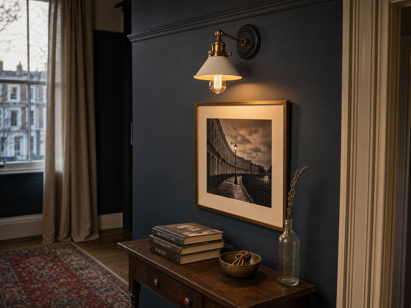 Bayswater white-cone-and-brass-collar wall sconce with Edison bulb mounted on a Hague Blue Edwardian hallway wall above a framed black-and-white photograph of a Bayswater crescent, antique walnut console table with stacked hardback books and brass keys in a small bronze bowl, Persian runner on oak floor, sash window onto stuccoed terrace at golden hour