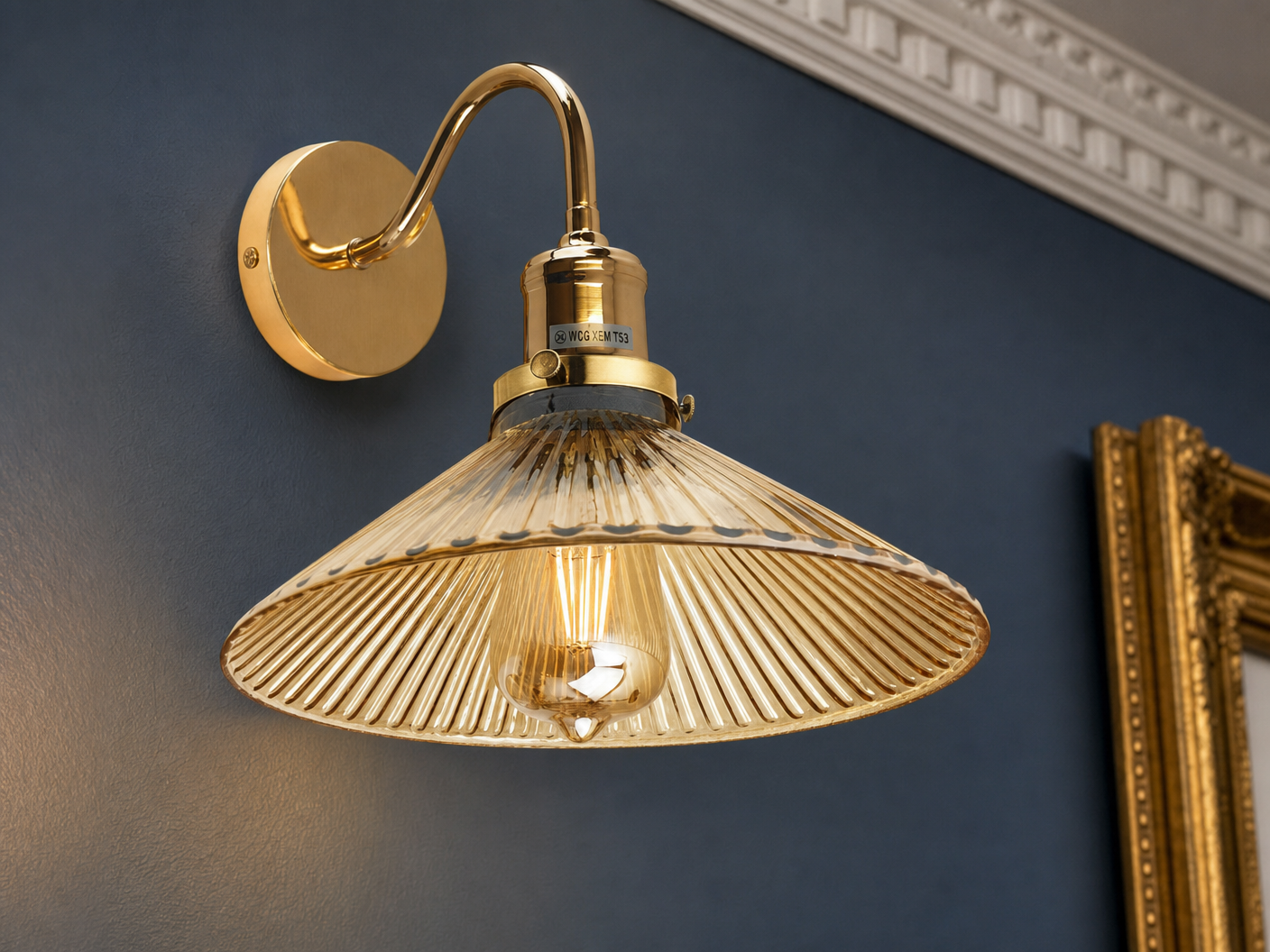 Belgravia detail close-up showing polished-brass S-curve gooseneck arm, brass collar with manufacturer label and small rivet detail, and wide amber-tinted fluted-glass coolie-hat shade with vertical flutes and Edison filament glowing inside, mounted on a Hague Blue plaster wall with Georgian dentil cornice