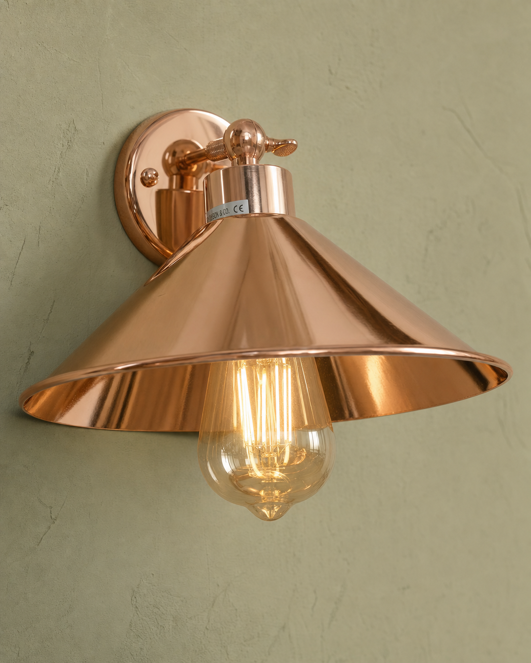 Cheltenham detail close-up showing highly polished mirror-bright rose-copper electroplated finish across the small disc wall plate, short straight stem with knurled adjustment knob/collar at the joint, flared coolie-hat shade with bell-mouth rim and polished interior, and clear amber-tinted ST64 Edison filament bulb hanging straight down through the centre, mounted on hand-plastered sage-green limewash