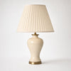 Salcombe table lamp on a soft warm-white background — full 56cm product view of the cream-glazed ceramic urn body on its antique-brass base plate beneath a tall classic empire-form shade in cream knife-pleated silk