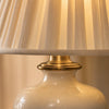 Detail of the Salcombe lamp — close-up of the cream glossy ceramic glaze with subtle kiln-fire variation, the polished antique-brass collar where the shade meets the urn, and the bottom edge of the cream knife-pleated silk shade catching warm light