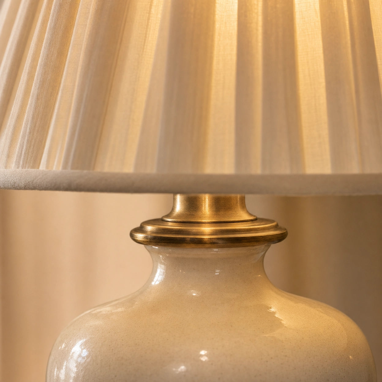 Detail of the Salcombe lamp — close-up of the cream glossy ceramic glaze with subtle kiln-fire variation, the polished antique-brass collar where the shade meets the urn, and the bottom edge of the cream knife-pleated silk shade catching warm light